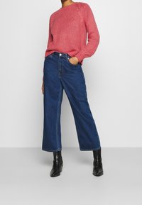 Růžový hrubě pletený svetr s žebrovanými detaily spojený s modrými vysoko pasovými džíny s širokými nohavicemi. Černé kotníkové boty dokončují outfit.