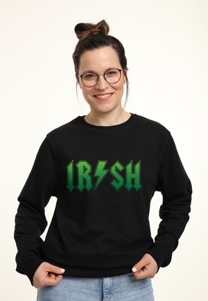 Smilende kvinde med briller og håret sat op i en knold iført en sort sweatshirt med grøn "IRISH" tekst og lyn, der holder i kanten.