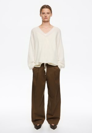 Woman standing with hands in pockets wearing oversized cream v-neck sweater, brown wide-leg trousers, and dark pointed shoes on white background.
