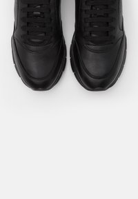 Svarta sportskor med en slät läderoverskikt, rund tå och texturerade skosnören. Yttersulan är av gummi med en rundad profil.