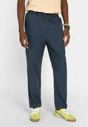 Hombre con pantalones casuales azul oscuro con cintura ajustable y zapatillas amarillas con cordones blancos sobre fondo liso.