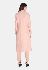 Cappotto lungo di colore rosa chiaro realizzato in un tessuto dalla consistenza liscia. Presenta una linea dritta e dettagli con bottoni sulle maniche.