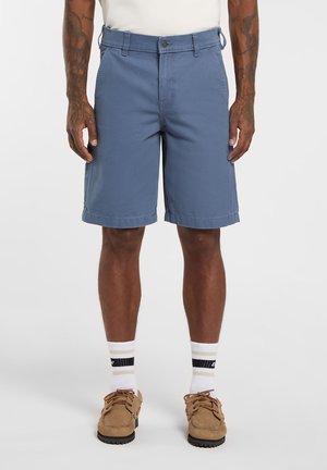 Blauwe katoenen korte broek met een rechte snit, riemlussen en knoopsluiting. Draagt de korte broek met beige schoenen, witte en zwarte gestreepte sokken.