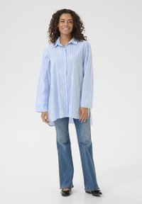 Svetlo modra srajca z dolgimi rokavi, belimi navpičnimi črtami in cvetličnimi vzorci, v kombinaciji z razširjenimi traperji in črnimi ravnimi čevlji.