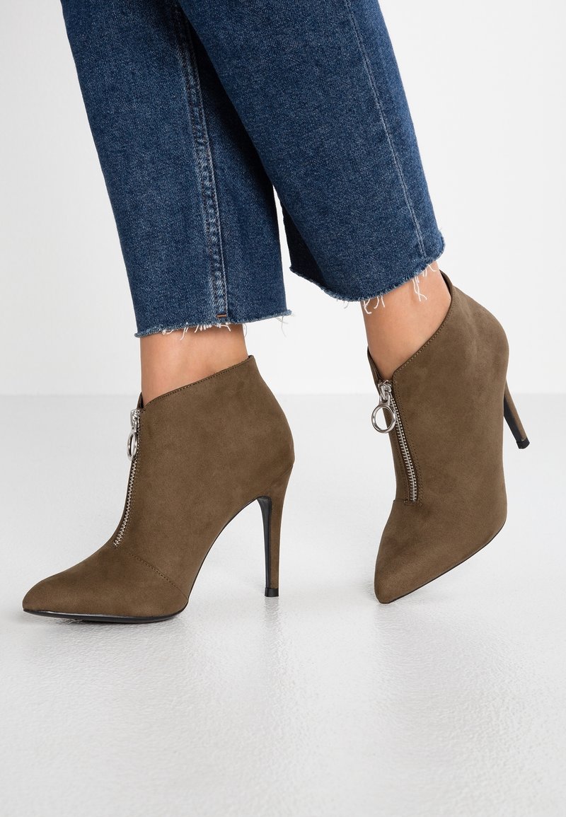 Bottines en daim marron à talons hauts avec fermeture éclair argentée à l'avant, portées avec un jean bleu à ourlet effiloché sur un sol clair.