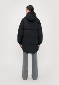 Svart boblejakke med hette, med et quiltet design og en lang, svakt buet kant. Stoffet ser teksturert ut, med elastiske mansjetter.