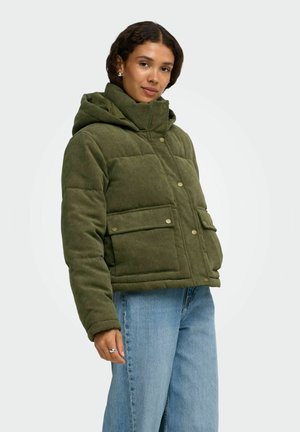 Femme portant une veste matelassée en velours côtelé verte avec de grandes poches devant et un jean bleu, debout devant un fond uni.