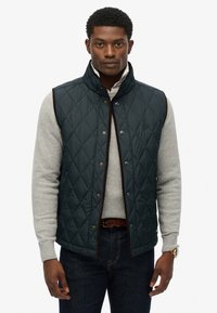 Quiltet navy vest med høj krave og brun kant, prydet med guld snapknapper og et subtilt logo, båret over en grå sweater.