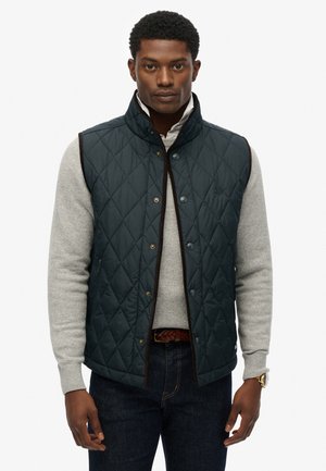 Quilted navy vest with a high collar and brown trim, featuring gold snap buttons and a subtle logo, worn over a gray sweater.