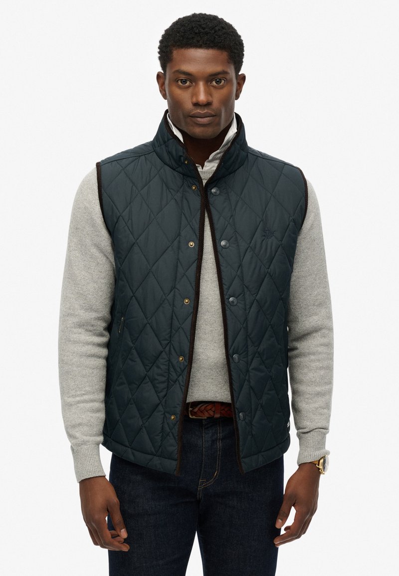 Quiltet navy vest med høj krave og brun kant, prydet med guld snapknapper og et subtilt logo, båret over en grå sweater.