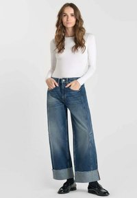 Donna con lunghi capelli mossi, che indossa una maglietta bianca a maniche lunghe, jeans larghi con risvolto, calzini neri e mocassini neri lucidi, in piedi con le mani in tasca.