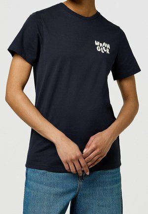 Osoba nosząca ciemnogranatowy t-shirt z krótkim rękawem z białym napisem "WRANGLER" na piersi i niebieskie dżinsy, ręce złączone z przodu.
