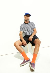 Camicia a righe con maniche corte, pantaloni corti neri, calzini arancioni, scarpe slip-on viola, indossando un cappello blu. Seduto su uno sgabello bianco contro uno sfondo bianco.