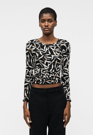 Γυναίκα που φοράει μαύρο τοπ με μακριά μανίκια και λευκά και μπεζ φυλλώδη μοτίβα, συνδυασμένο με μαύρο ψηλόμεσο παντελόνι, στέκεται μπροστά σε απλό φόντο.