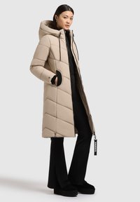 Dlhý béžový prešívaný kabát s kapucňou, s diagonálnymi prešívanými sekciami, čiernymi detailmi na vreckách a zipsom. Štylizovaný s čiernymi úzkymi nohavicami.
