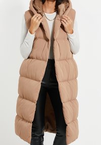 Longue doudoune sans manches couleur beige, dotée d'une capuche, de boutons-pression et d'une texture matelassée, portée sur un haut blanc et un pantalon noir.