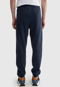 Muž v tmavě modrých teplákových kalhotách a modrých teniskách stojí s rukama uvolněnými podél těla před jednobarevným pozadím.