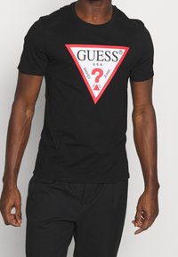 Camiseta negra de manga corta con un gran gráfico de triángulo rojo y blanco con el diseño de "GUESS U.S.A." y un signo de interrogación en el centro.