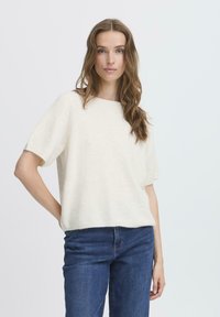 Lichtcrème korte mouwen trui met een zachte textuur en een rechte snit, gedragen met donkerblauwe jeans, handen in de zakken.
