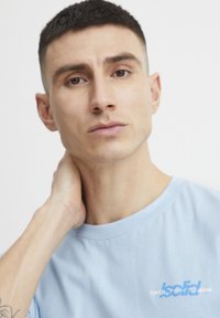 Lyseblå bomull T-skjorte med rund hals og korte ermer, med et trykt logo i hvitt og blått på brystet.