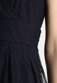 Robe bleu marine en tissu texturé avec motifs en relief. Conception plissée et superposition en tissu transparent visibles, mettant en valeur des détails complexes.