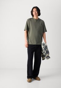 T-shirt olive à manches courtes avec une poche poitrine, associé à un pantalon large bleu foncé et une chemise à carreaux par-dessus. Chaussures beiges.