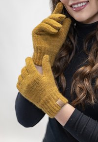 Pletené rukavice hořčicové barvy vyrobené z měkkého materiálu. Mají žebrované manžety a kožený akcent s logem na zápěstí.
