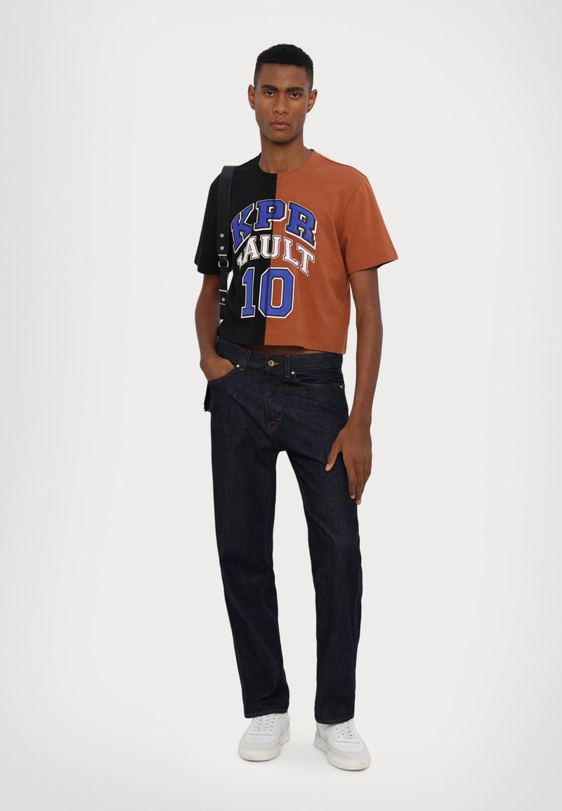 Cropped short-sleeve shirt with black and brown halves, featuring blue lettering; paired with dark jeans and white sneakers.