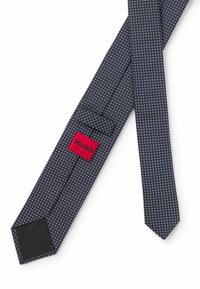 Marinblå slips med geometriska mönster, med en framträdande röd etikett som läser "HUGO" och "MADE IN ITALY". Slipsen har en spetsig ände och en klassisk stil.
