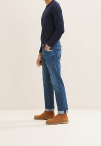 Homme portant un jean bleu retroussé, des bottines marron à lacets et une chemise sombre à manches longues, debout devant un fond uni.