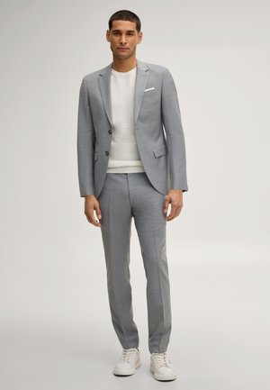 Junger Mann, der einen hellgrauen Anzug, einen weißen strukturierten Pullover und weiße Sneakers auf einfarbig hellem Hintergrund trägt