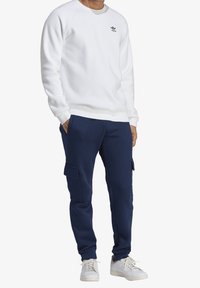 Bílá mikina s žebrovanými manžetami a lemem, doplněná tmavě modrými cargo kalhotami s bočními kapsami a bílými teniskami.