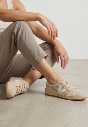 Person siddende på gulvet iført beige bukser og beige sneakers med lysebrune såler, hænder hvilende på knæ mod ensfarvet baggrund.