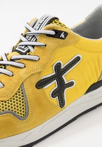 Żółte trampki wykonane z zamszu i siateczki, z czarnymi akcentami, białymi sznurówkami i charakterystycznym wzorem logo na boku.