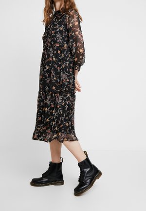 Kobieta w czarnej kwiecistej sukience midi z przezroczystymi rękawami i czarnych sznurowanych botkach, stojąca na jednolitym jasnym tle.