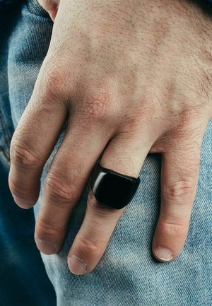 Black rectangular ring on a hand, featuring a smooth finish. The hand is positioned against light blue denim fabric.