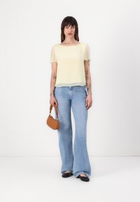 Haut transparent jaune clair à manches courtes, associé à un jean bleu évasé taille haute et un petit sac à main marron. Des ballerines noires complètent le look.