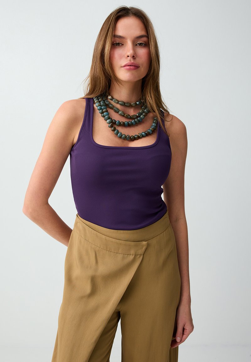 Jeune femme portant un top violet sans manches, un collier superposé de perles vertes et un pantalon marron taille haute à jambes larges sur fond uni.