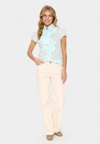 Camicia menta chiara con dettaglio a volant sul collo, maniche corte; abbinata a pantaloni beige chiaro a gamba larga e ballerine marroni con borchie.