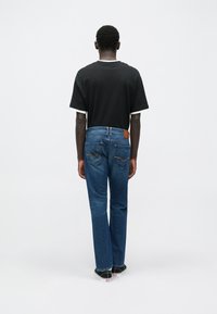 Fekete póló fehér szegéllyel, kék egyenes szárú farmernadrág és fekete cipő fehér talppal. Hátulnézetből látható.