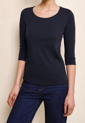 Kobieta ubrana w dopasowany czarny top o długości rękawa do połowy przedramienia, w połączeniu z ciemnoniebieskimi dżinsami, stojąca na tle neutralnego beżowego tła.