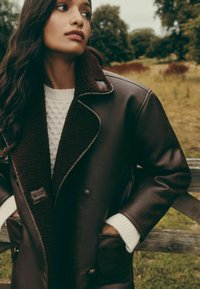 Pruun nahktagi tume, karvase kraega, esitaskute ja nööpidega, kombineeritud tekstuurse valge kampsuniga.