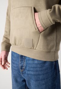 Hoodie bege com uma textura suave, apresentando um bolso canguru e um emblema texturizado. Combinado com jeans azuis.