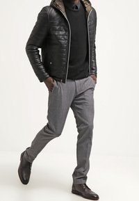 Giacca nera in pelle trapuntata con cappuccio, indossata sopra un maglione scuro. Pantaloni eleganti grigi e scarpe in pelle marrone completano l'outfit.