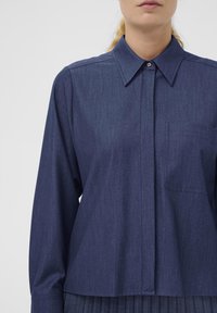 Granatowa koszula z długim rękawem, wykonana z fakturowanej tkaniny. Posiada kołnierzyk, długie rękawy i kieszonkę na piersi. Prosty design z minimalnymi detalami.