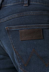 Jeans de mezclilla azul oscuro con parche de logo de cuero marrón de Wrangler en el bolsillo trasero, con costuras naranjas y un patrón de zigzag.