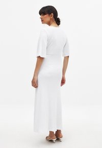 Robe maxi blanche texturée avec un corsage ajusté et des manches évasées, présentant un décolleté rond et une silhouette lisse et fluide.