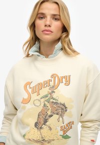 Sweatshirt crema con grafica di cowboy in arancione e verde, con testo "SuperDry" e "SANTA ROSA", colletto e polsini a coste. Tessuto leggermente texturizzato.