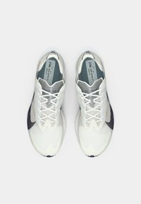 O pereche de pantofi de alergare Nike VaporFly albi cu logo-uri Swoosh negre văzuți de sus pe un fundal simplu deschis.