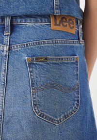 Blå denimjeans med en brun läderlapp med "Lee"-logotyp och en liten svart etikett på bakfickan. Synliga sömdetaljer.